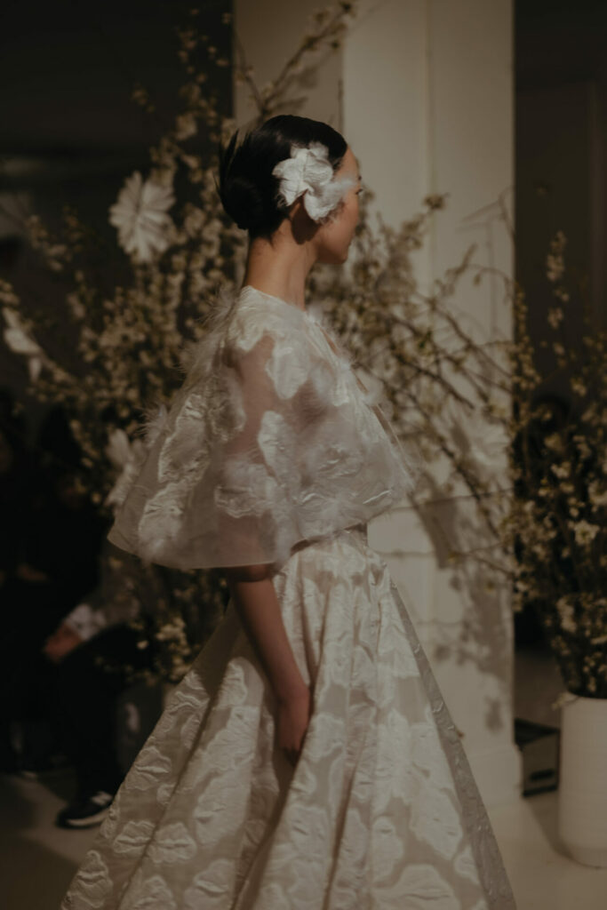 Bride in sheer floral dress, flowers blurred behind.