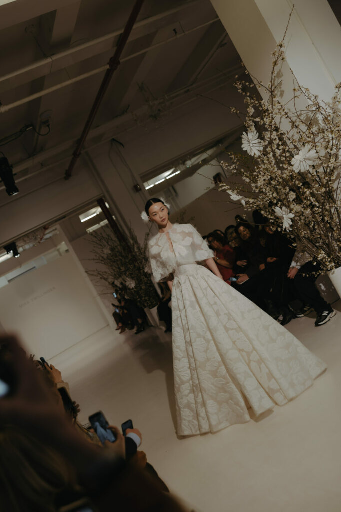 Model in floral Clea wedding dress with cape on runway.