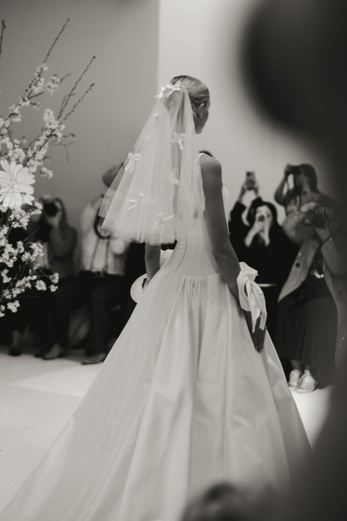 Elegant bride in Clea dress passes guests; timeless, formal scene.
