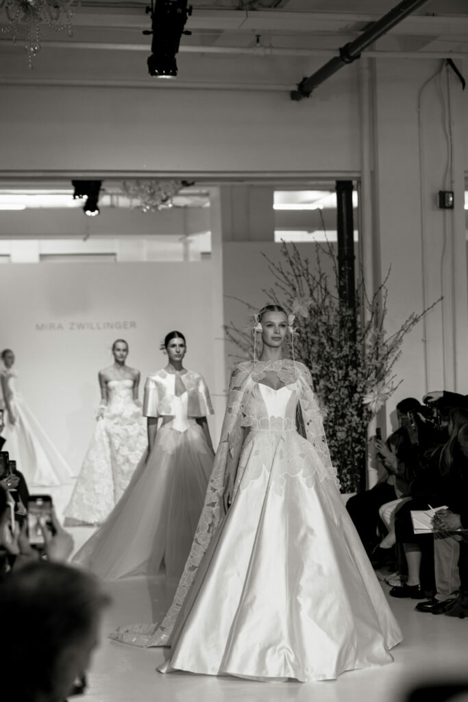 Mira Zwillinger SS2027 bridal models walk runway in lace couture gowns at floral New York Bridal Fashion Week venue.