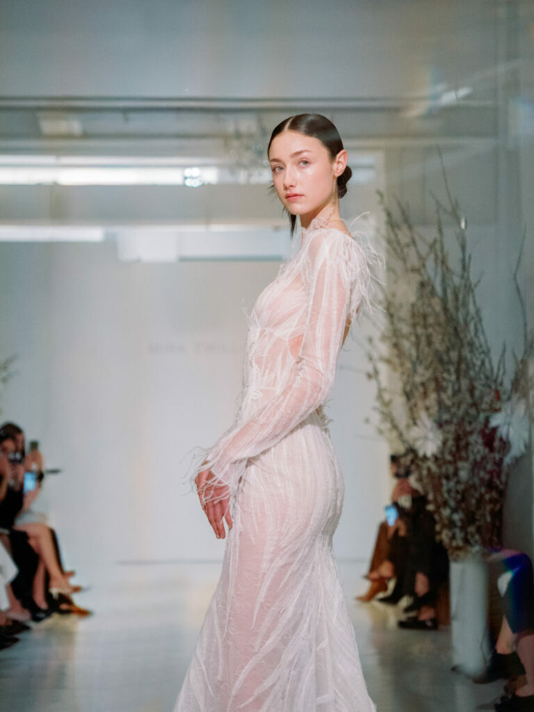Model in sheer white gown on runway, florals behind.