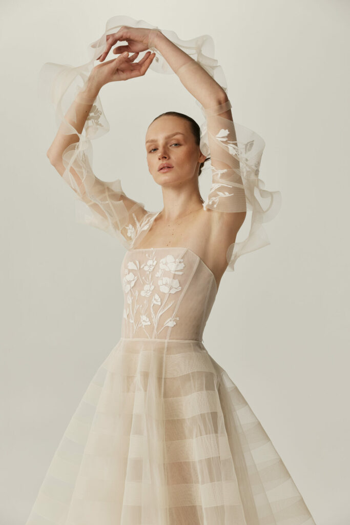 Woman in sheer Clea wedding dress, arms raised.