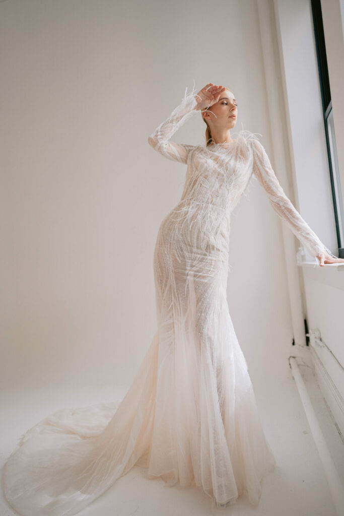 Woman in sheer lace gown gazes out window.
