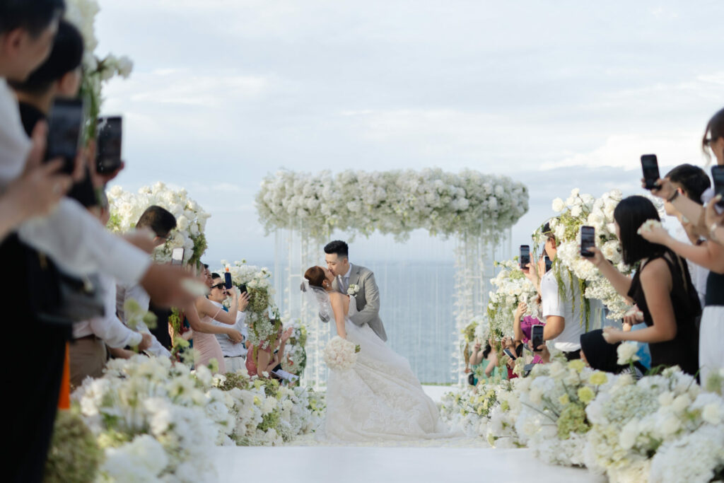 Bride and groom kiss under white floral arch, ocean behind.
