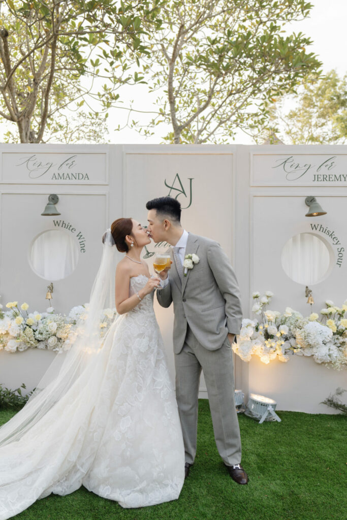 Bride and groom smile, holding drinks by floral backdrop.