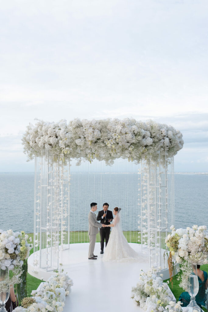 Bride, groom, and officiant under white floral arch by ocean.