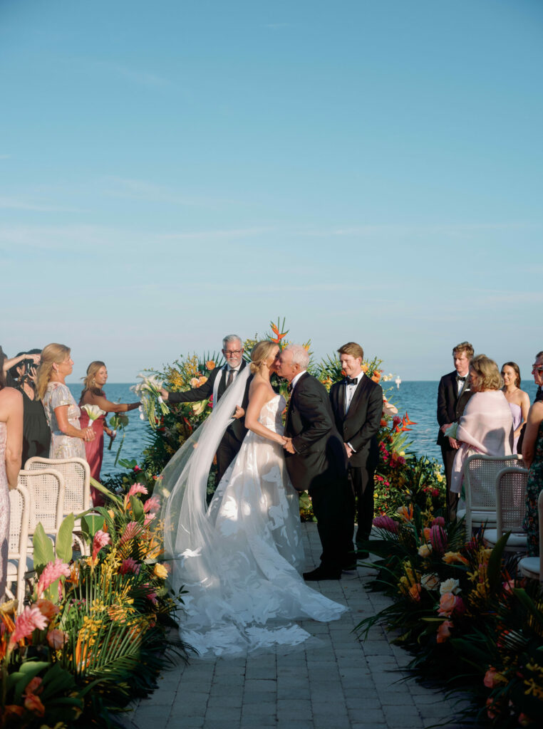 Bride and groom kiss at colorful oceanfront ceremony.