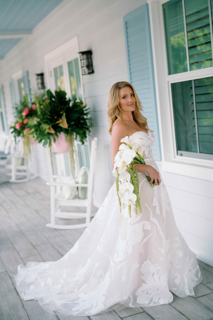 Bride in floral gown holding orchid bouquet on porch.