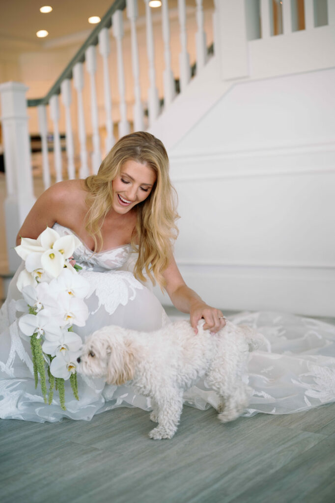 Smiling bride kneels petting fluffy dog by stairs.