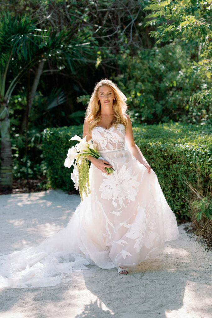 Bride in sheer floral gown walking with bouquet outdoors.