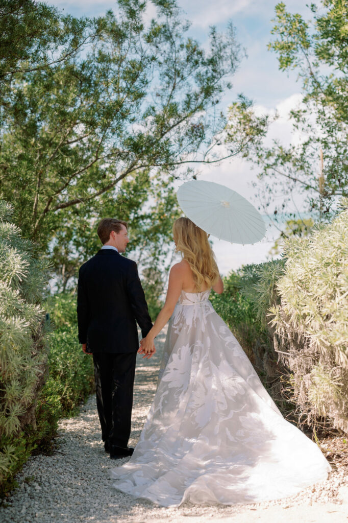 Bride in couture with parasol walks garden path with groom.