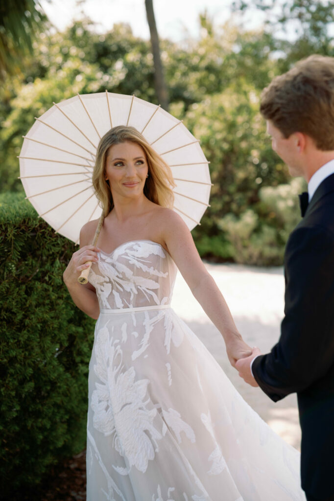 Bride in couture, white parasol, hand-in-hand outdoors.