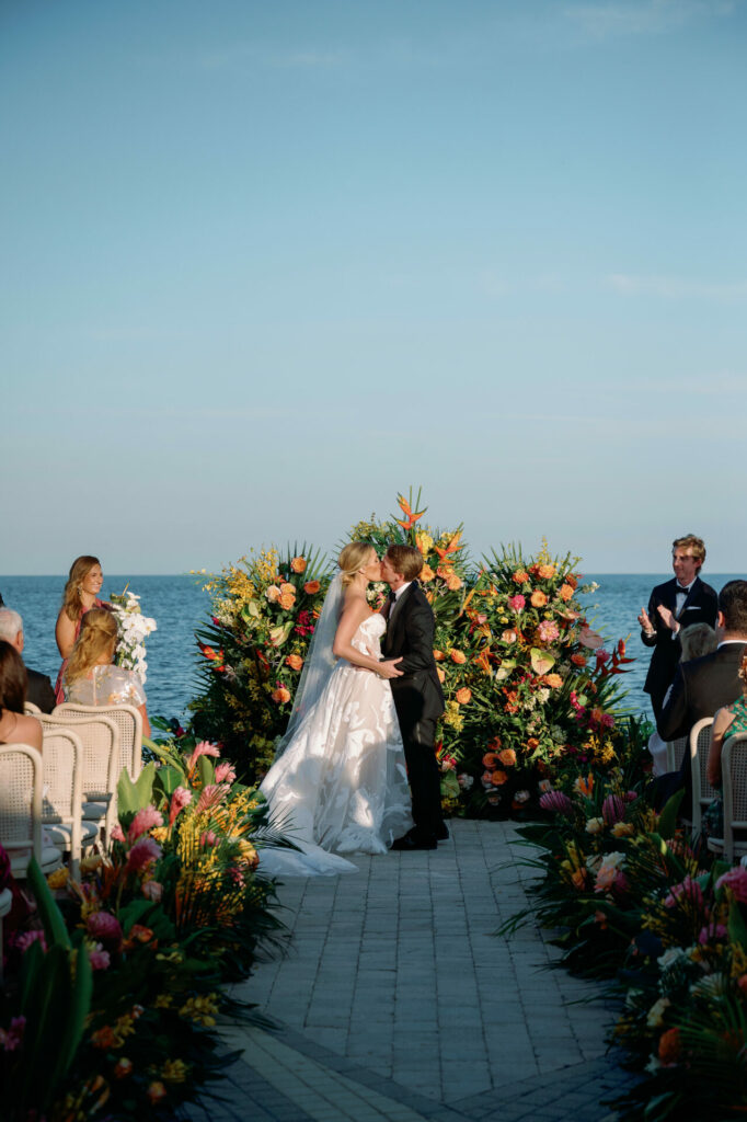 Bride and groom kiss by ocean amid tropical blooms.