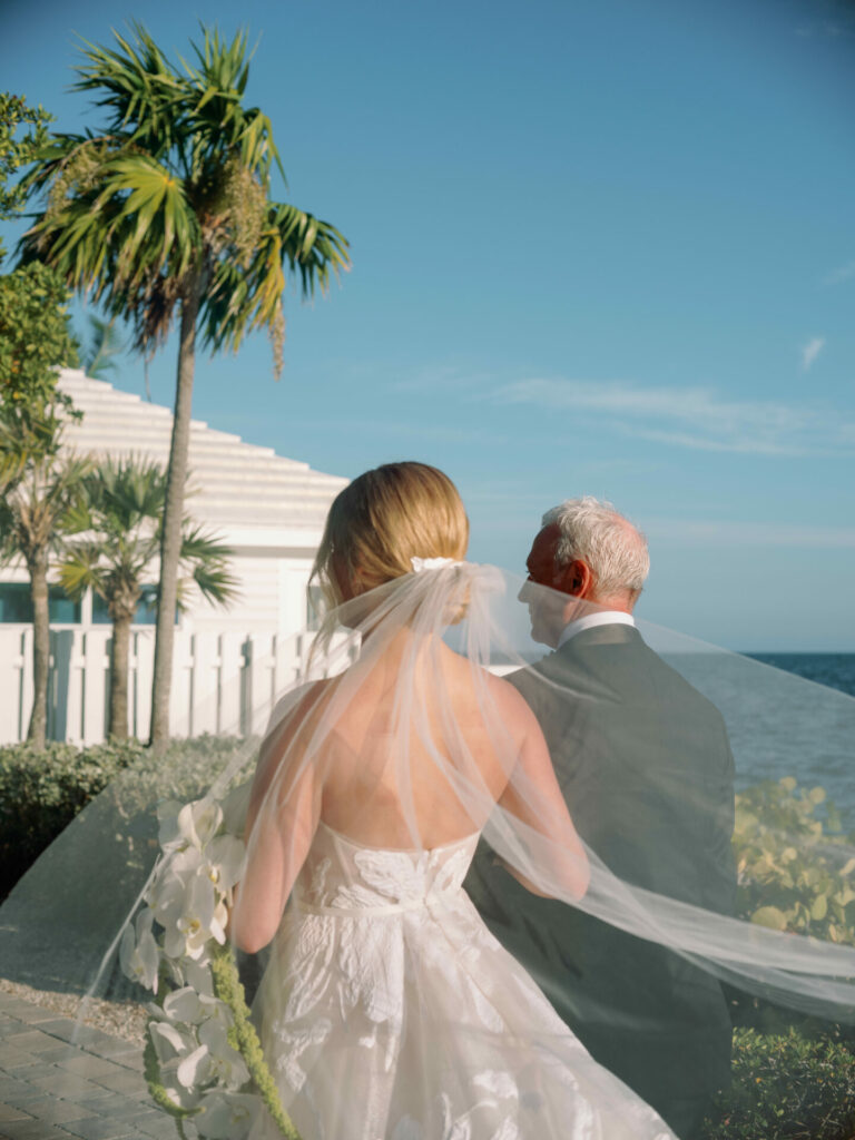 Bride in luxury gown and veil walks with older man outdoors.
