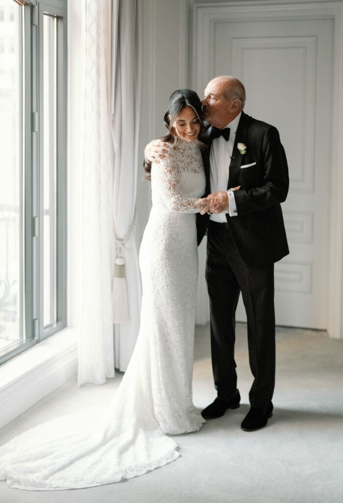 Bride in long-sleeved gown hugged by older man.