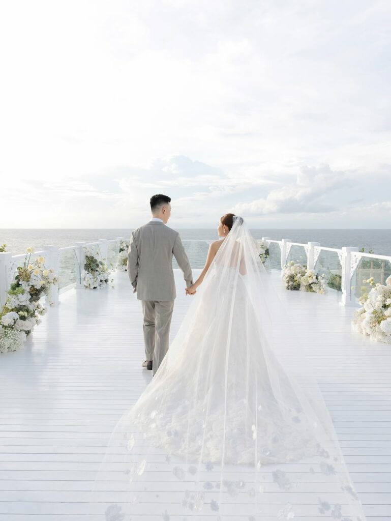 Bride and groom on floral deck, ocean view, sunny sky.