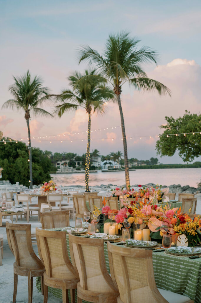 Chic sunset dining by water, flowers, candles, string lights.