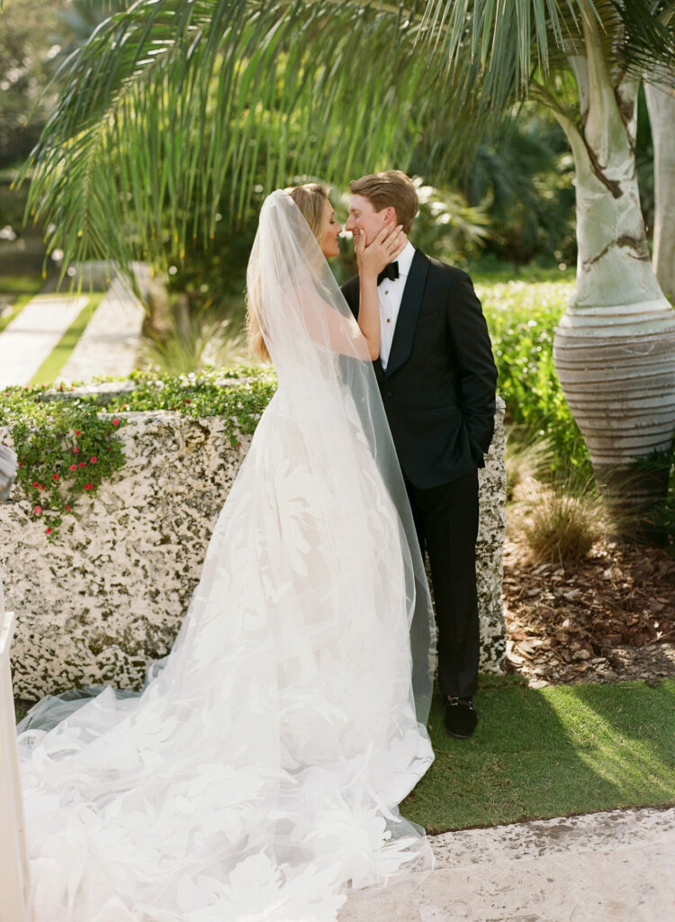 Bride in couture touches groom’s face outdoors.