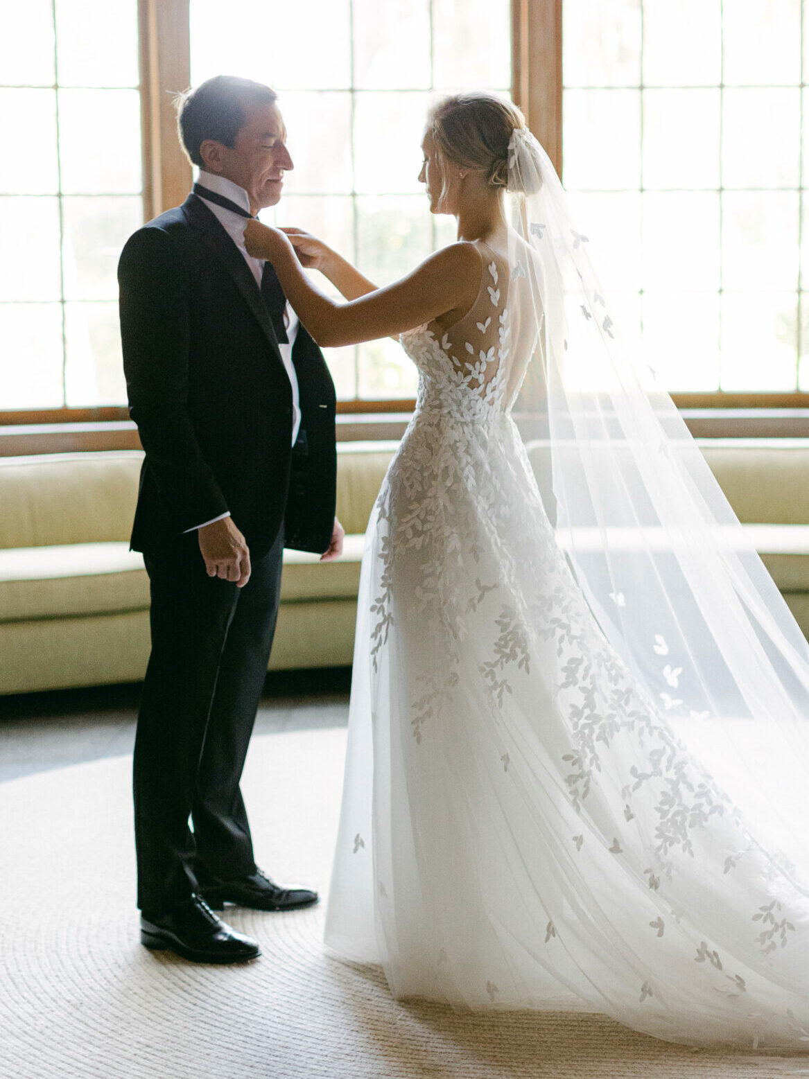 Bride fixes groom's bow tie in sunlit room.
