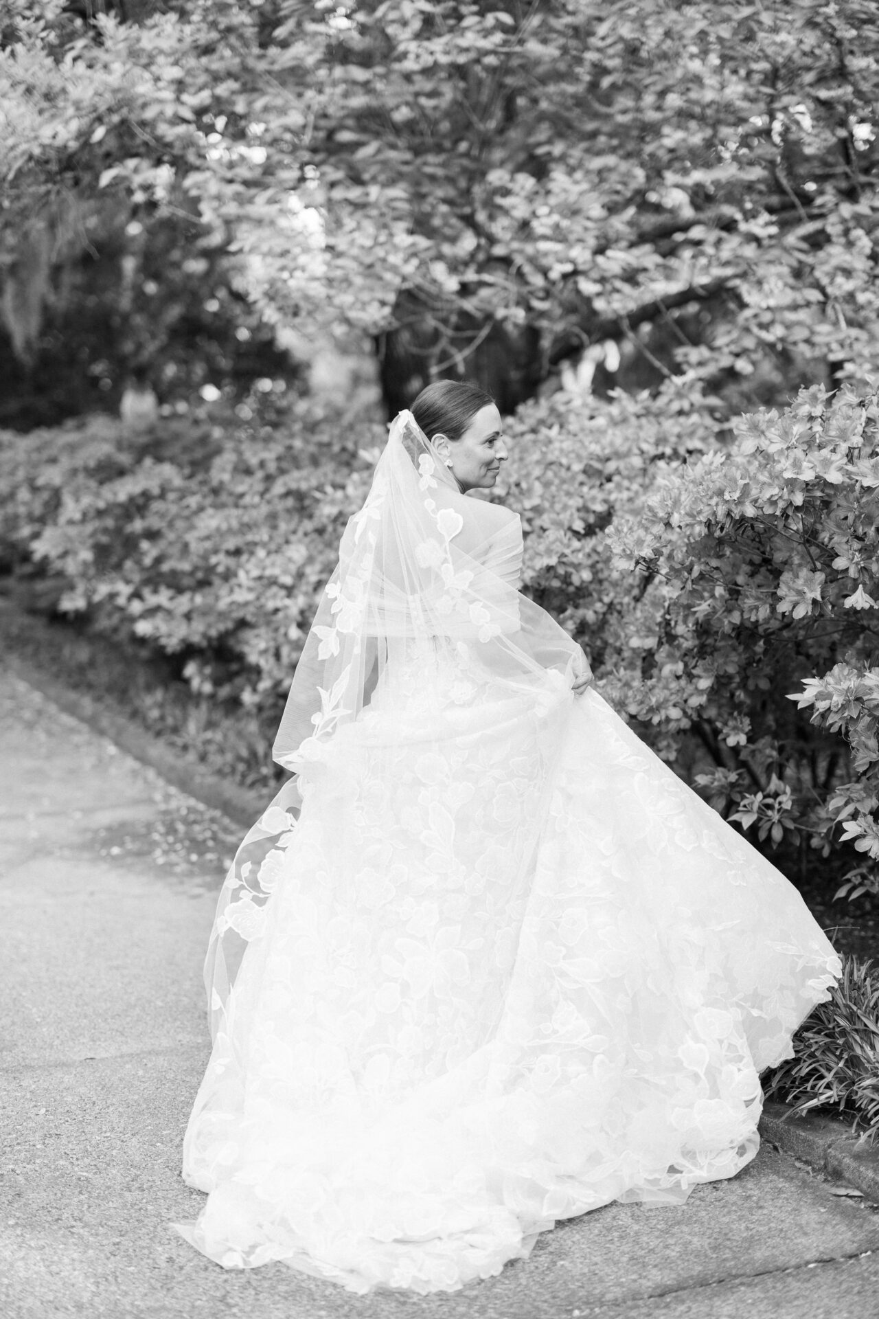 Smiling bride in floral dress walks garden path, monochrome.
