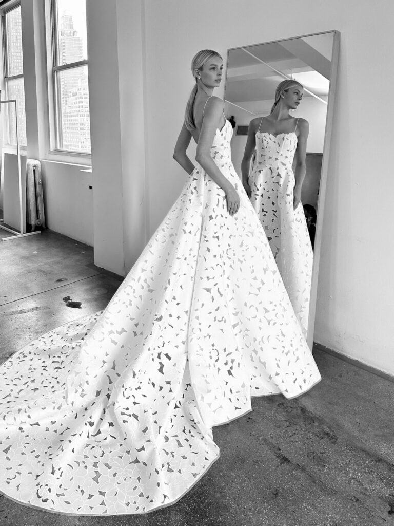 Woman in patterned white gown by mirror, reflection visible.