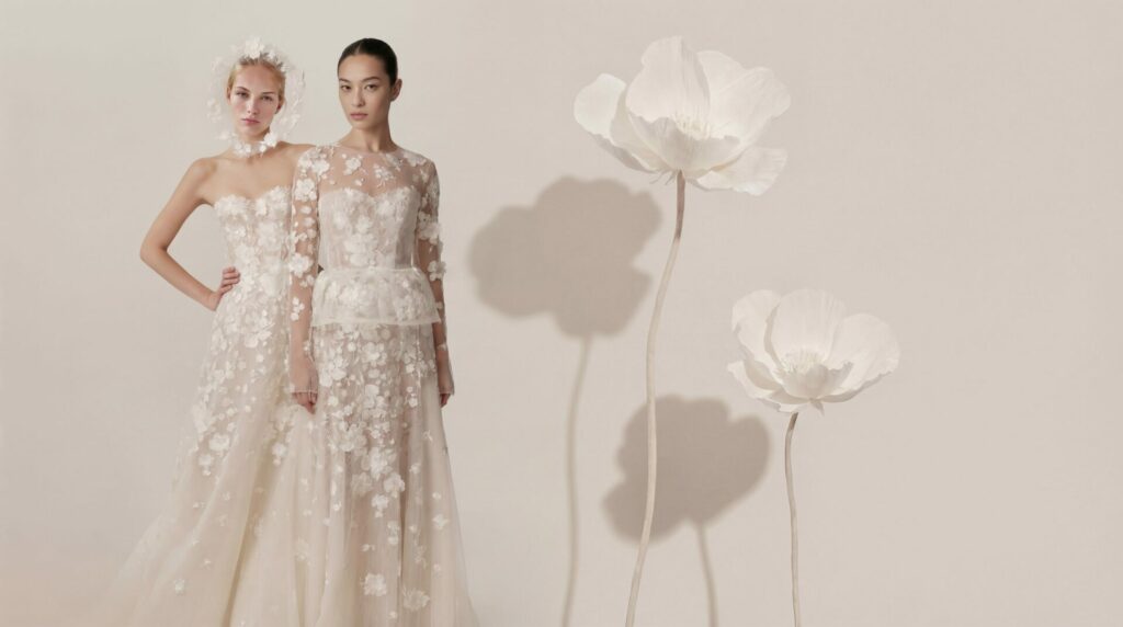 Two women in white gowns stand before floral backdrop.