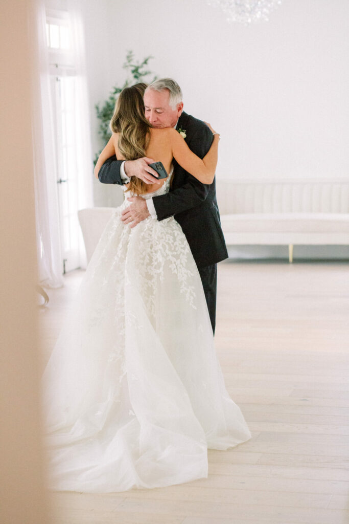 Bride in Clea dress hugs older man in bright room.