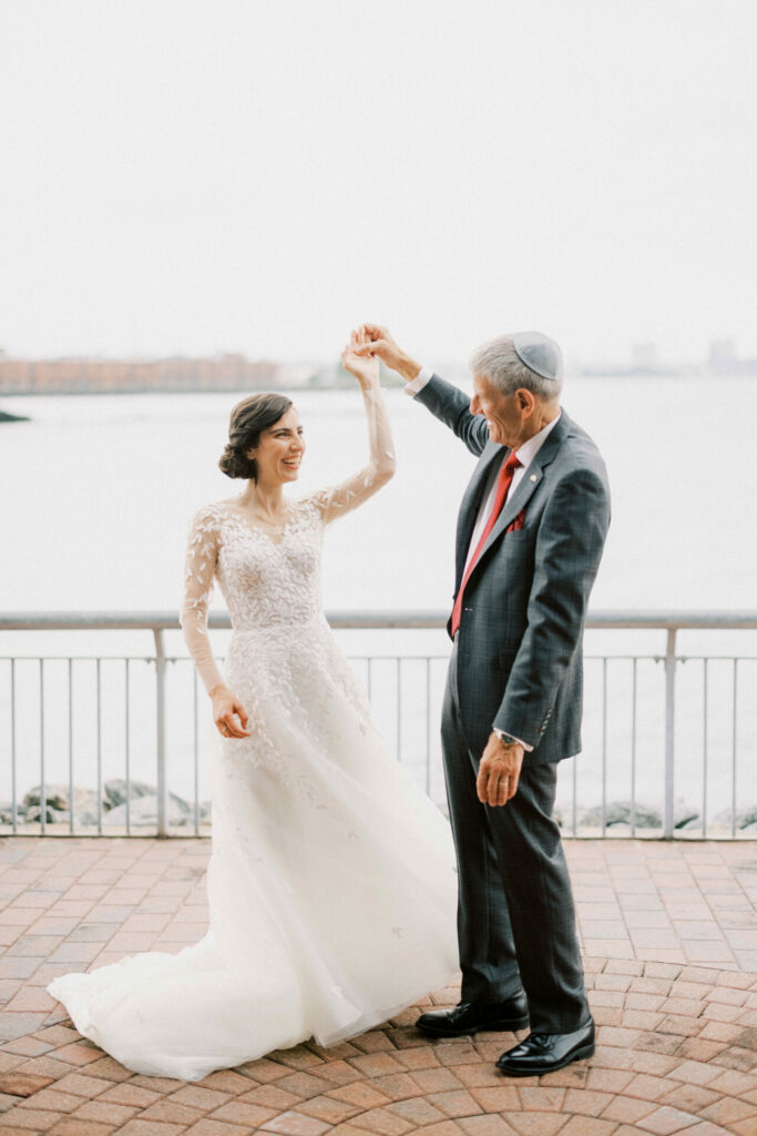 Bride and man in kippah dance by waterfront.