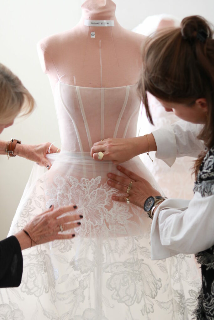 Two people adjust sheer floral lace dress on mannequin.