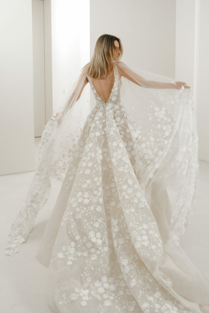 Blonde woman in lace gown holds sheer train.
