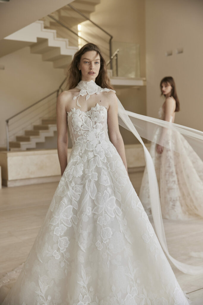 Bride in lace gown with bow; blurred figure behind.