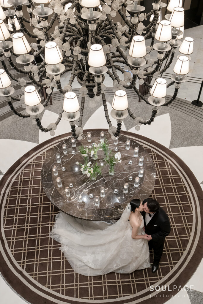 Bride and groom kiss beside marble table, chandelier overhead.