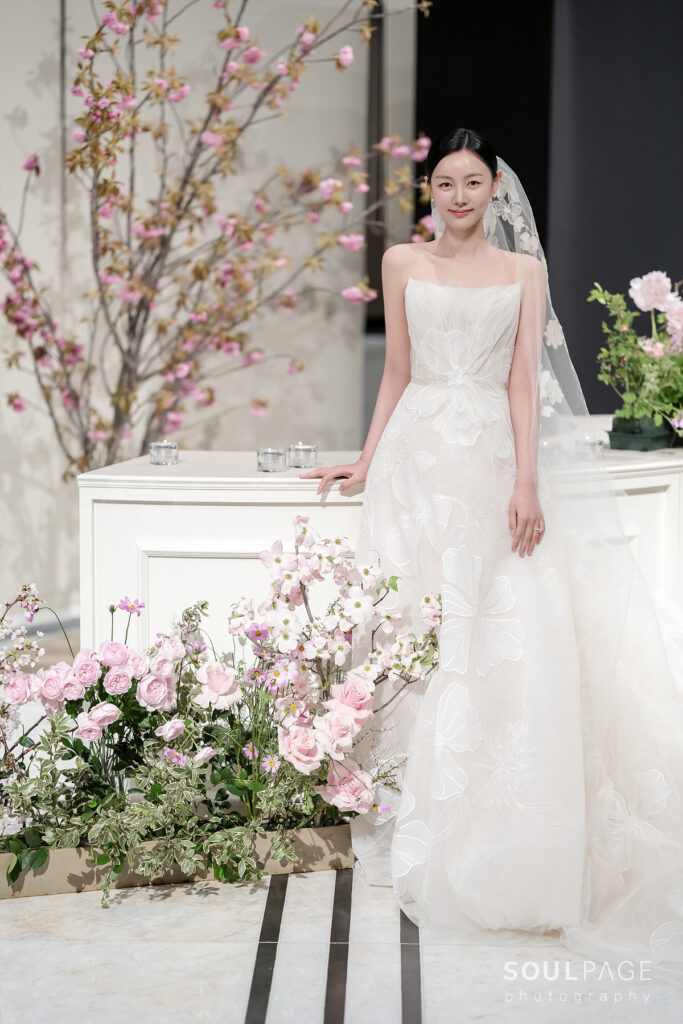 Bride in white dress by pink flowers, smiling.