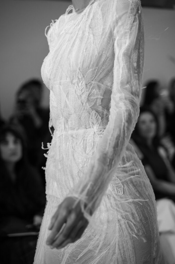 Model in sheer white lace dress on runway, monochrome.
