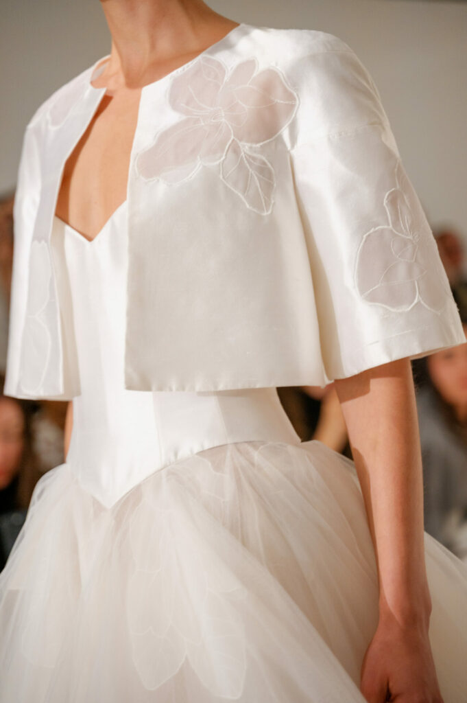 Model in Clea wedding dress with floral jacket; blurred crowd background.
