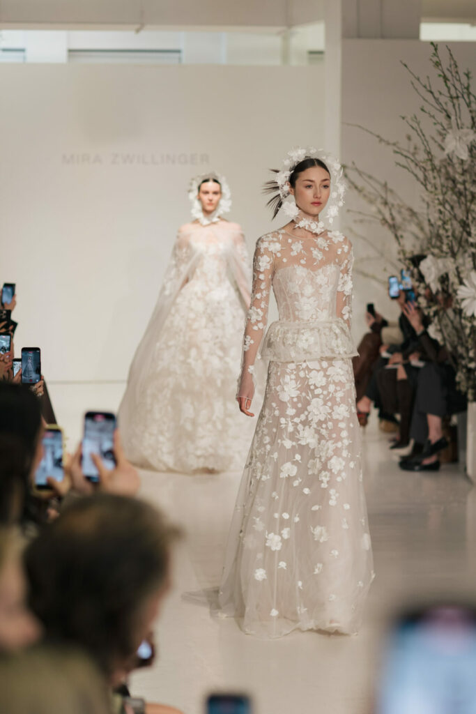 Two models in white floral bridal gowns on runway.