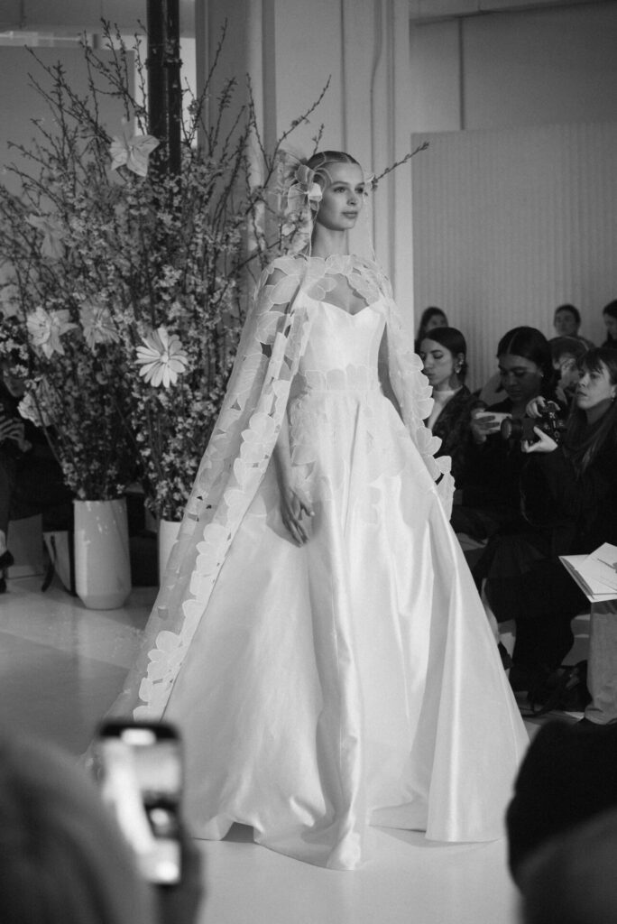 Model in white gown walks amid flowers, spectators.