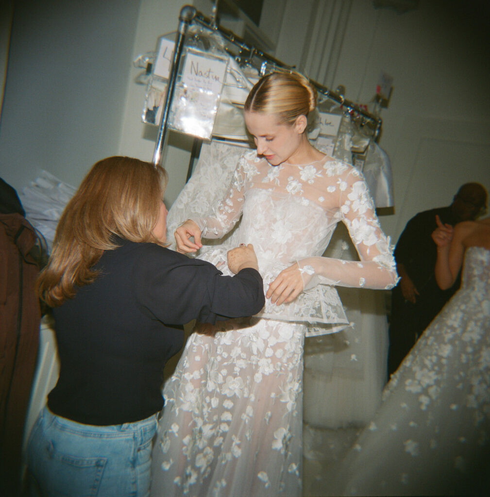 Designer fits model’s sheer floral gown backstage; others nearby.