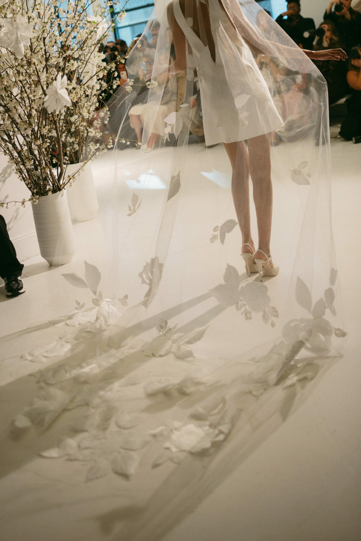 Bride in short white dress, sheer floral veil, flowers nearby.