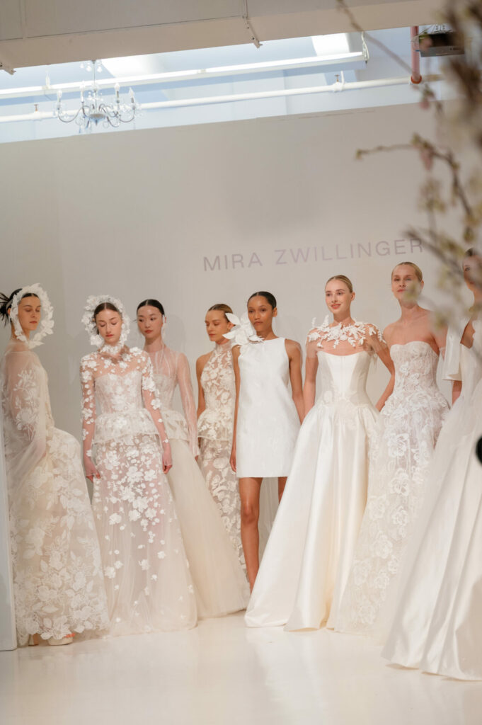 Seven models in white dresses, Mira Zwillinger background.