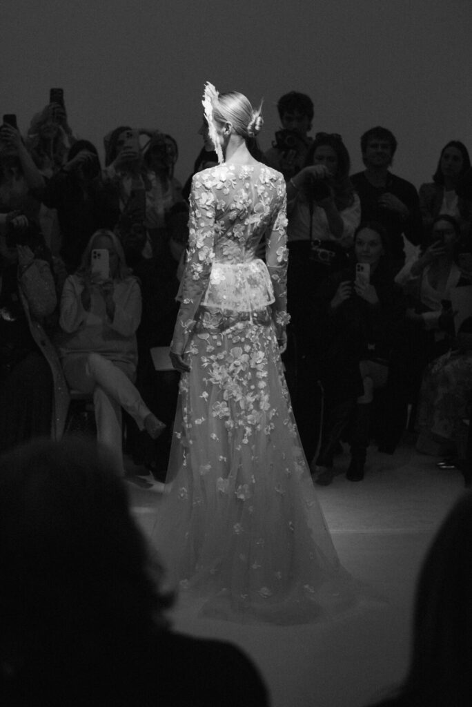Model in floral sheer gown with wing headpiece, runway, black and white.