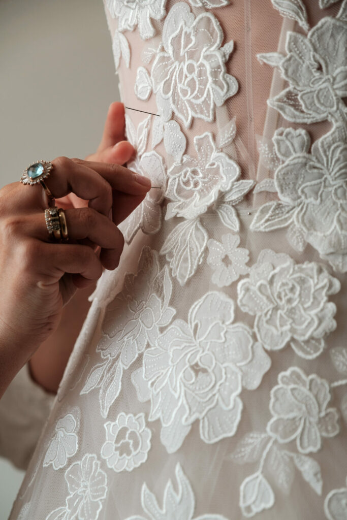 Hands buttoning lacy floral wedding dress, rings visible.