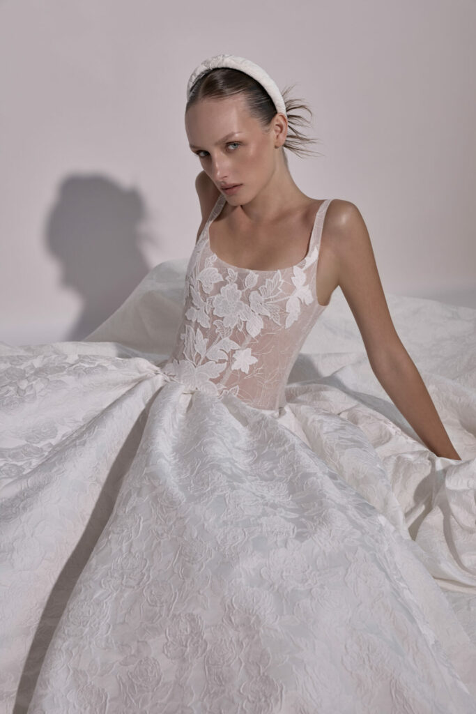 Woman in lace gown and headband sits, neutral expression.