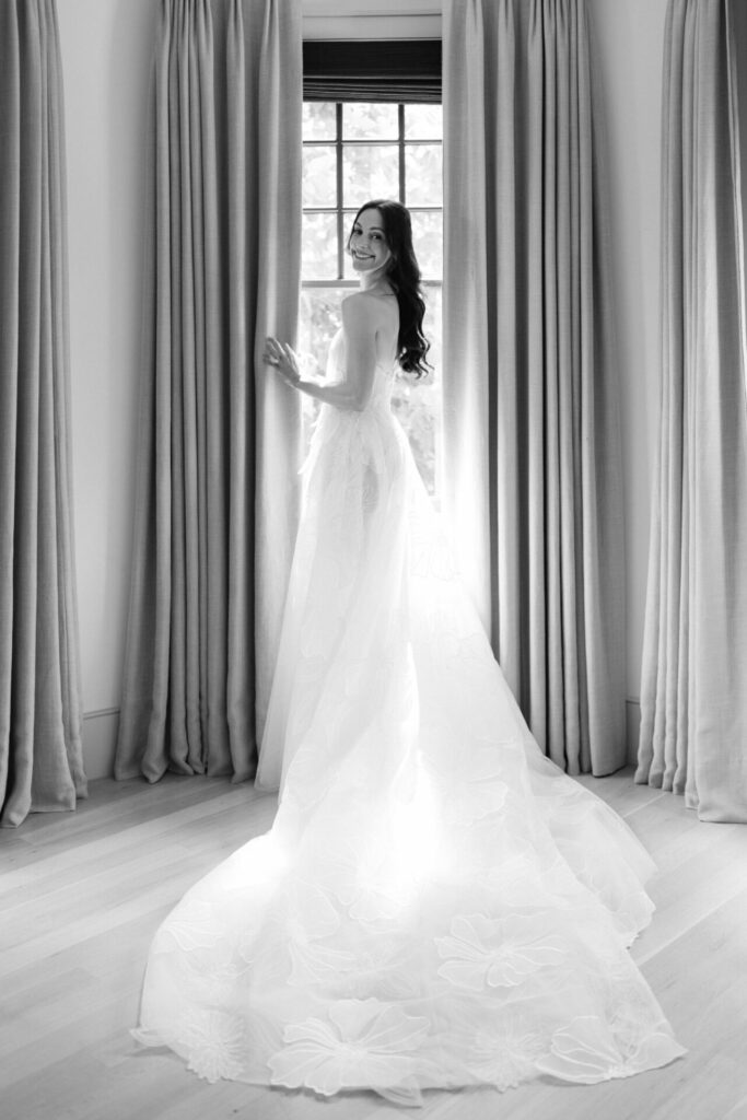 Bride in strapless gown smiles by window, black and white.
