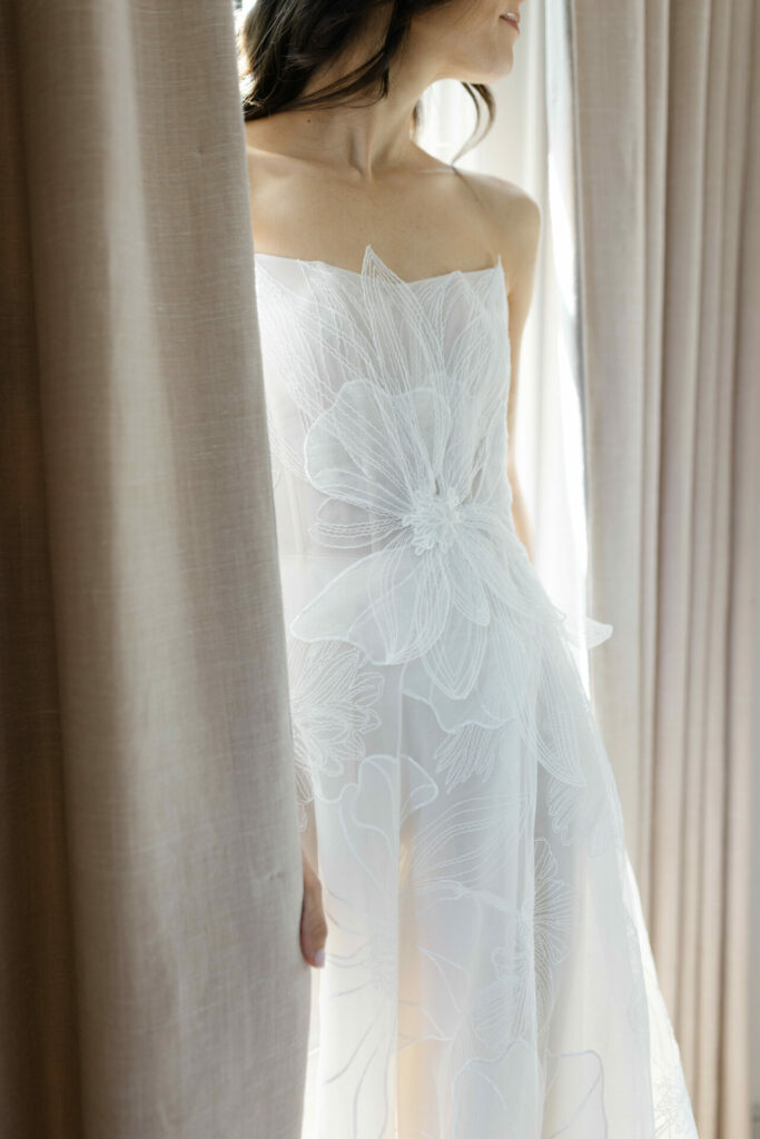 Woman in sheer white floral dress between beige curtains.