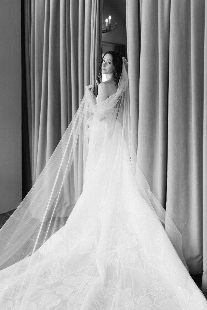 Bride in flowing gown stands between curtains, wistful.