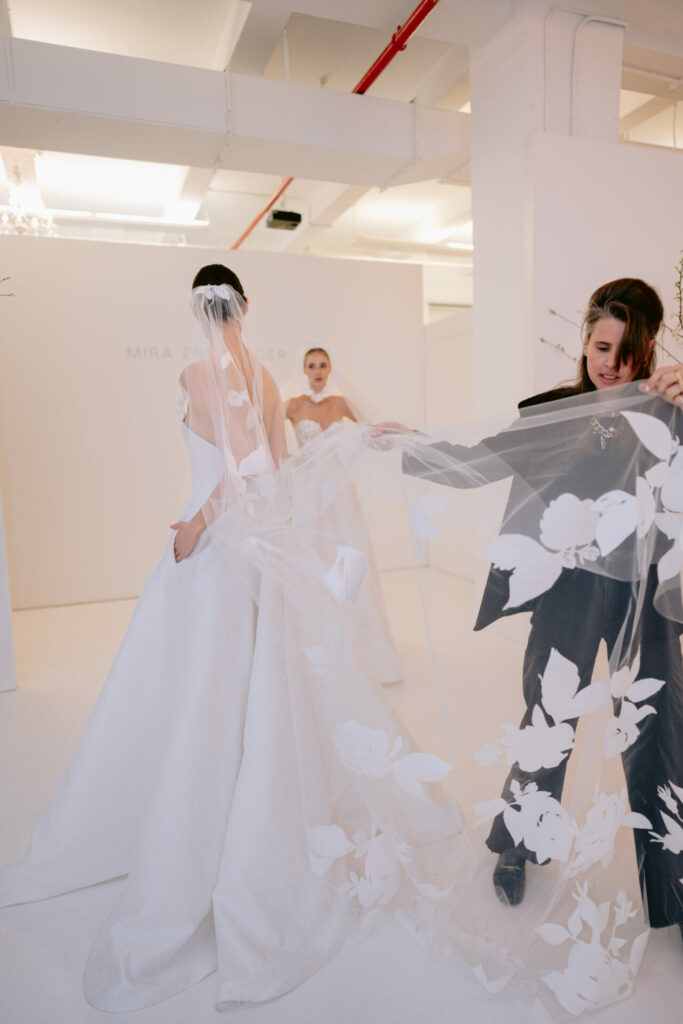 Two brides indoors; one’s floral veil is adjusted.