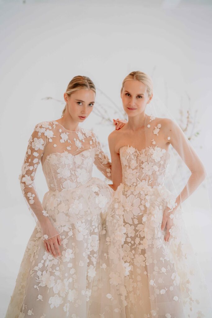 Two women in white floral wedding gowns pose together.