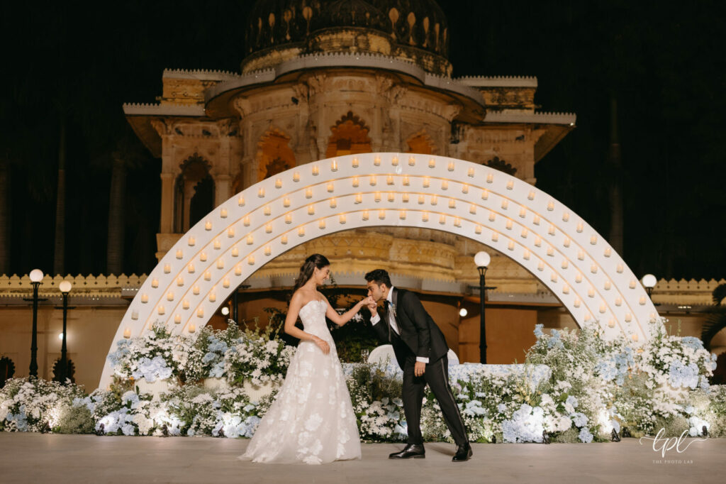 Groom kisses bride’s hand under lit wedding arch at night.