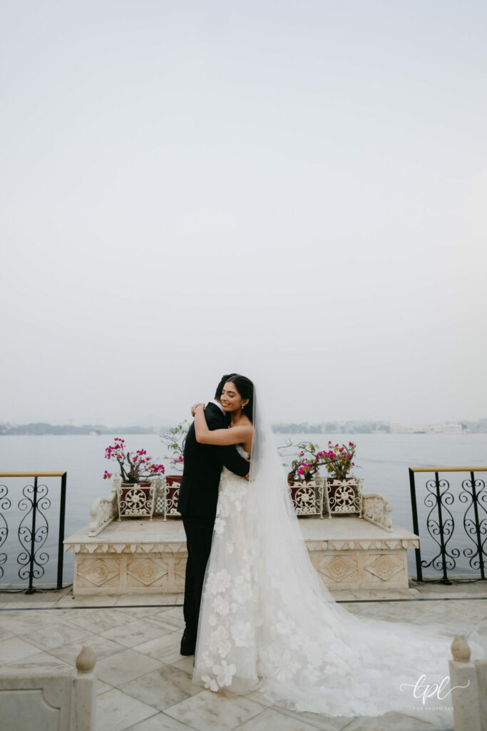Bride in floral gown embraces groom on lakeside terrace.